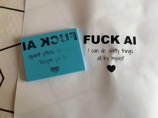 Une feuille avec imprimé « FUCK AI I can do shitty things all by myself » et un cœur, et une petite plaque de gomme à graver, avec le même motif dessus.